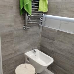 Wwww - Bidet wiszący pod grzejnikiem drabinkowym z zielonymi i niebieskimi ręcznikami, obok stoi mały, okrągły stołek na tle szarych płytek imitujących drewno.