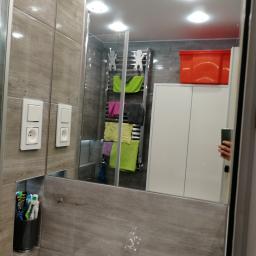 Wwww - Wnętrze łazienki z lustrem, w którym odbija się grzejnik na ręczniki z kolorowymi ręcznikami, biała szafka i czerwony pojemnik na górze. Na ścianie widoczne dwa gniazdka elektryczne i pojemnik...