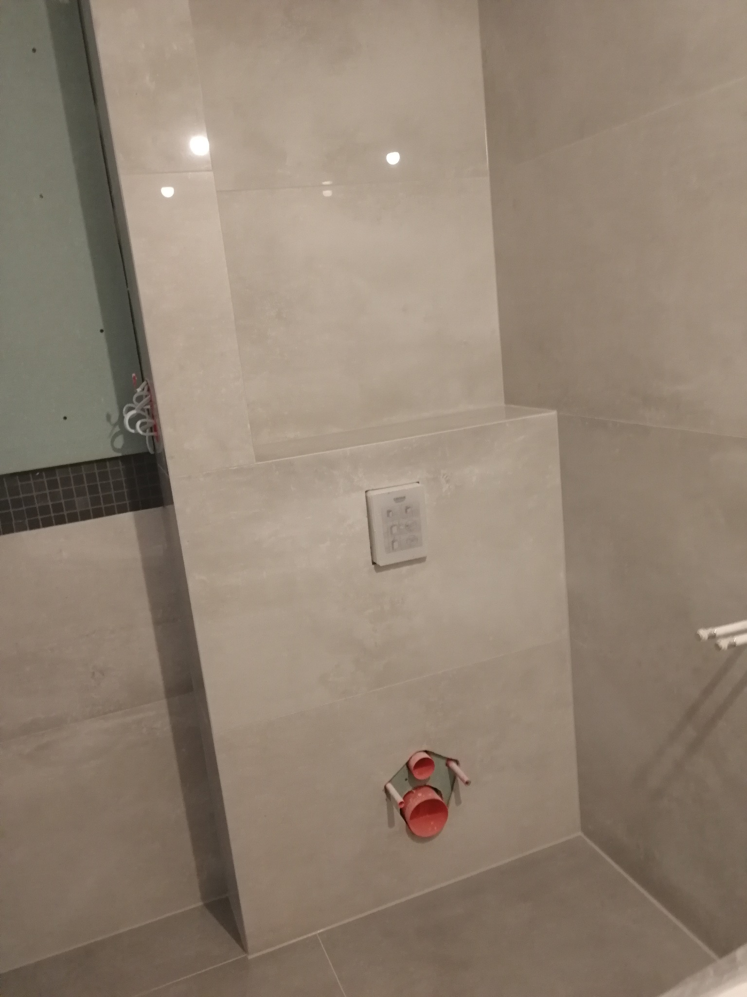 Szara łazienka w trakcie prac wykończeniowych, widoczne płytki, zabudowa geberitu z przyciskiem i instalacja wodna z czerwonymi rurami.