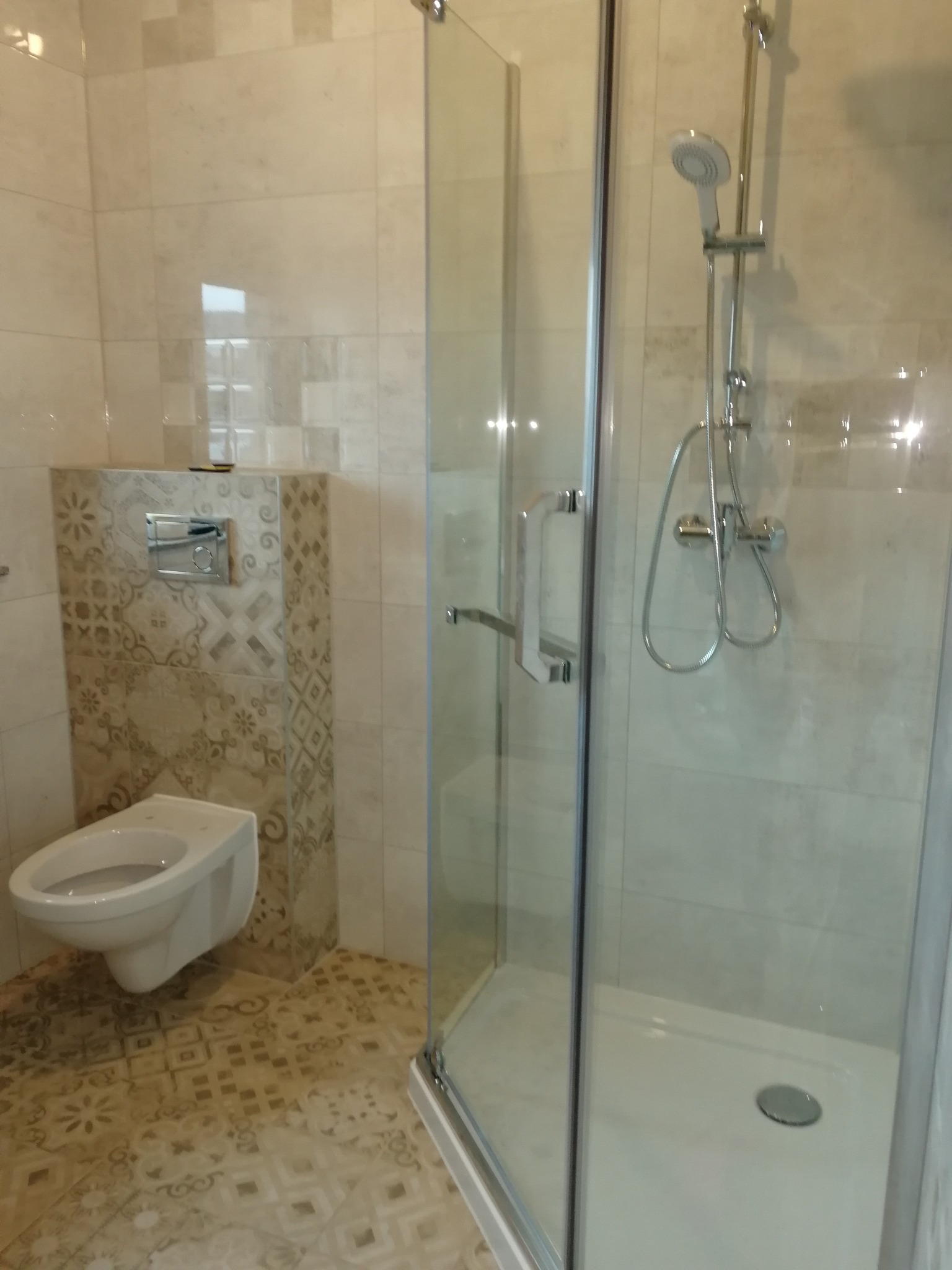 Wykończona łazienka z kabiną prysznicową ze szklanymi drzwiami, wiszącą toaletą z ozdobną obudową oraz podłogą z wzorzystych płytek.