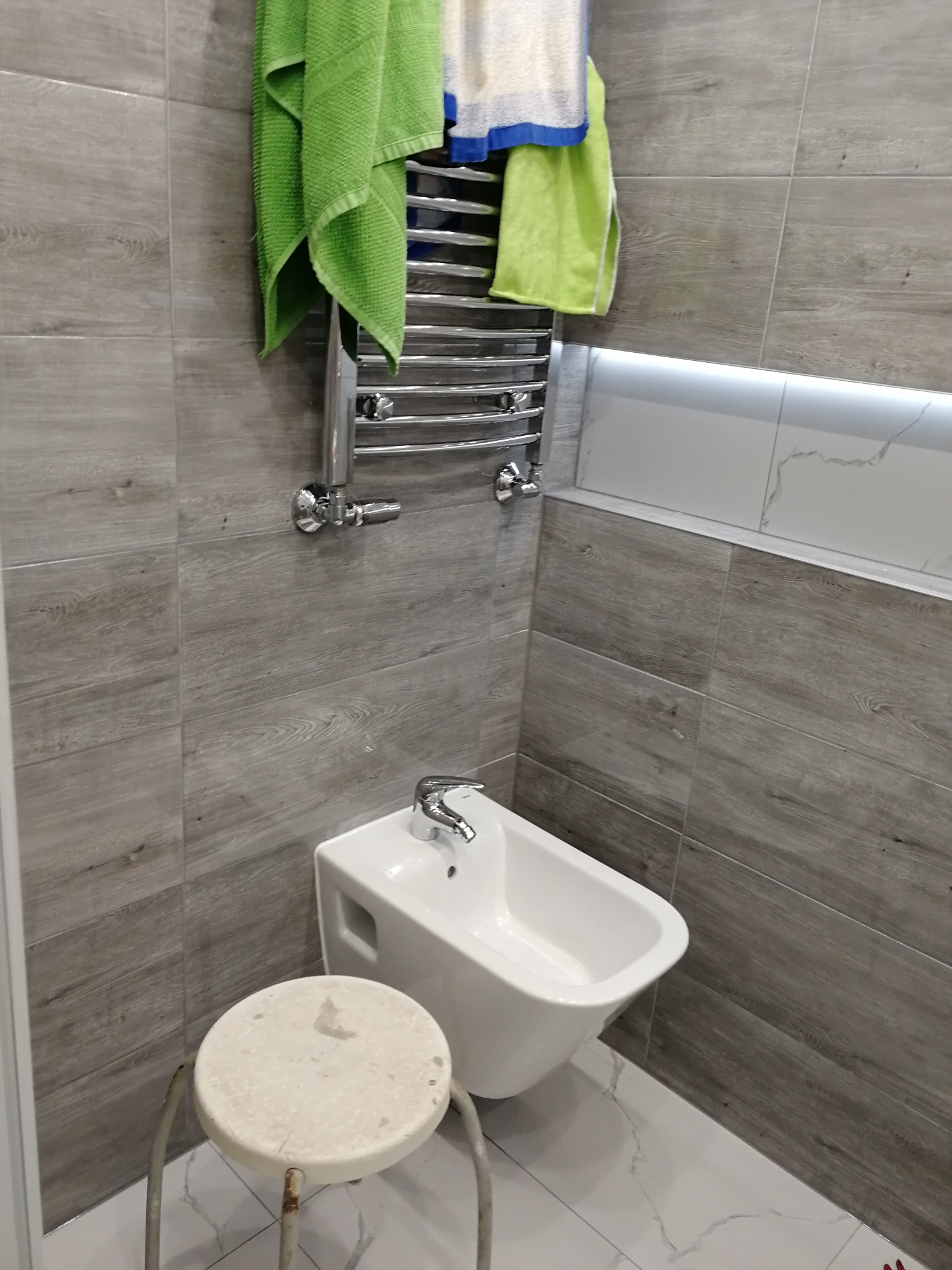 Bidet wiszący pod grzejnikiem drabinkowym z zielonymi i niebieskimi ręcznikami, obok stoi mały, okrągły stołek na tle szarych płytek imitujących drewno.