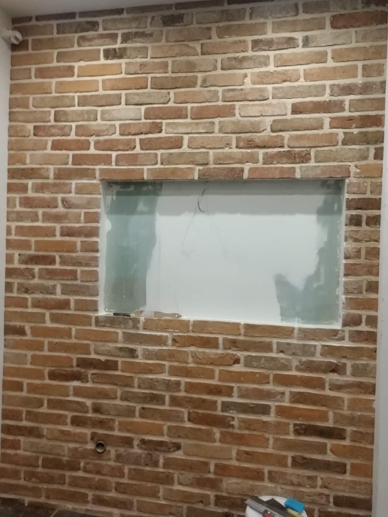 Ściana z cegły klinkierowej z wnęką przygotowaną do malowania, widoczne narzędzia malarskie na dole kadru.