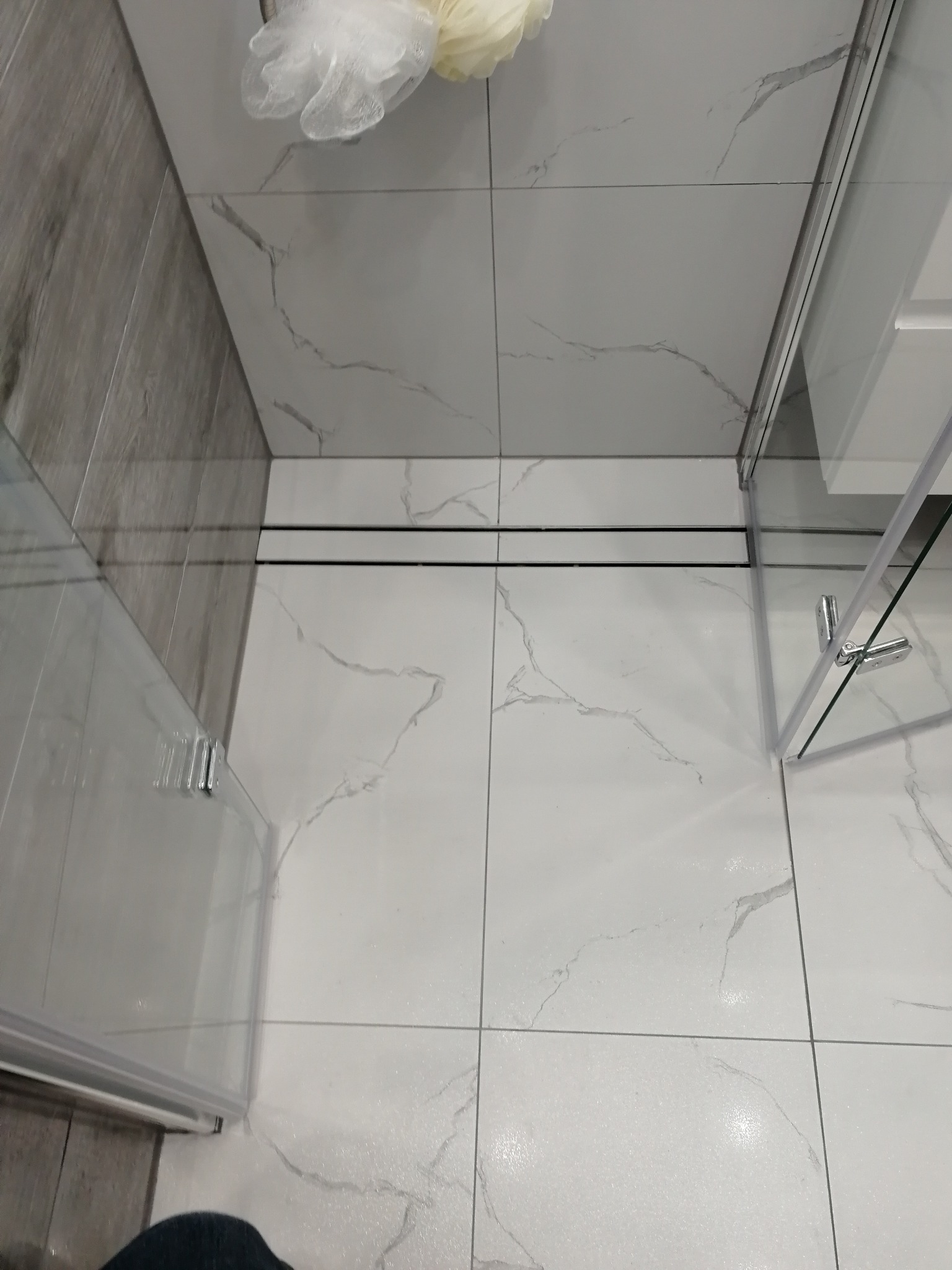 Białe płytki podłogowe z szarym wzorem marmuru, czarny odpływ liniowy i fragment kabiny prysznicowej z przezroczystego szkła.