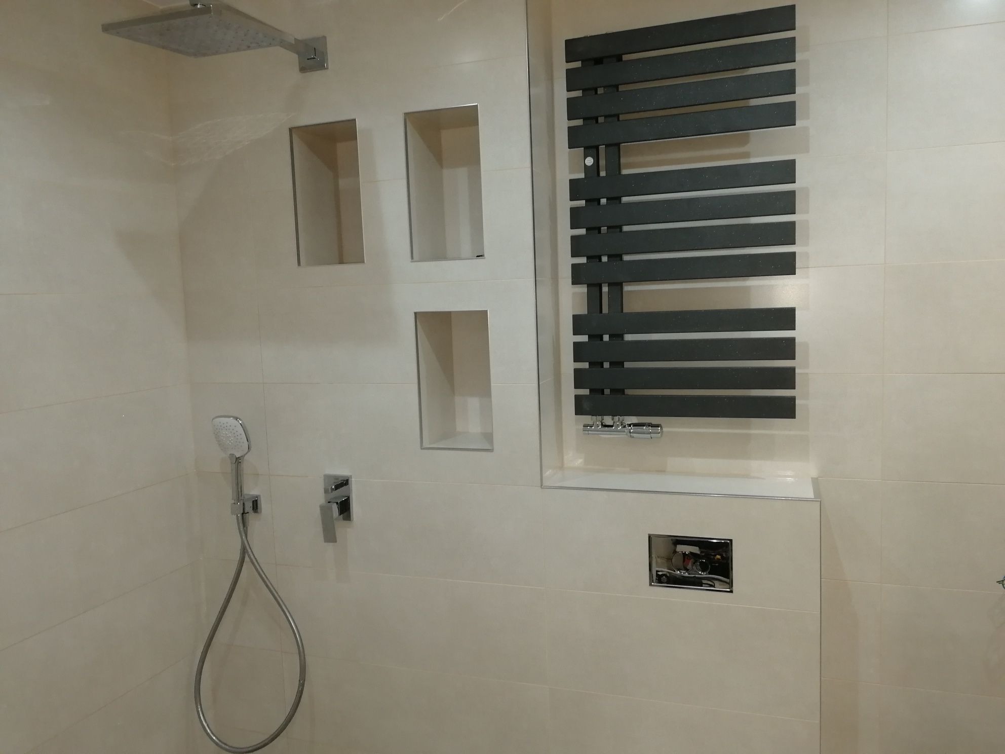 Wykończona łazienka z beżowymi płytkami, chromowaną armaturą, grzejnikiem drabinkowym w kolorze antracytowym i wnękami w ścianie.