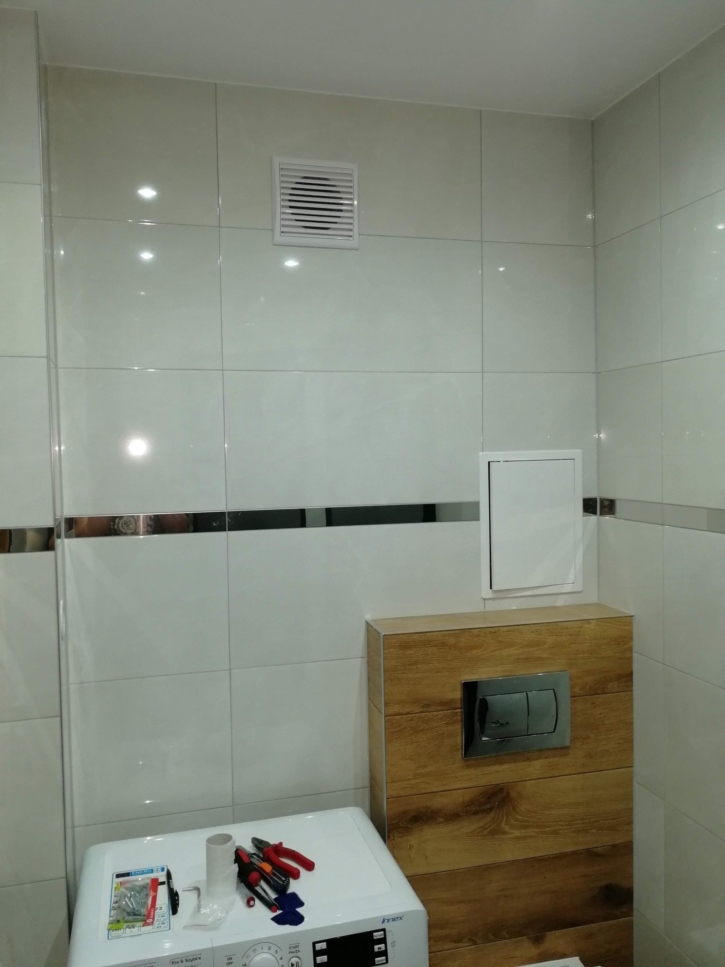 Błyszczące, białe płytki w łazience z dekoracyjnym pasem ze stali nierdzewnej, wentylator i ukryta spłuczka WC, narzędzia i śruby na pralce.