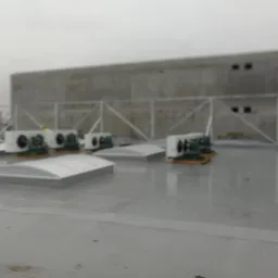 Szary, płaski dach budynku przemysłowego z zainstalowanymi klimatyzatorami przemysłowymi i świetlikami dachowymi, zabezpieczony metalową balustradą na tle betonowej ściany.