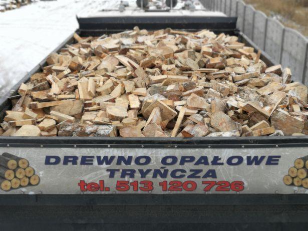 Przyczepa pełna pociętego drewna opałowego, widoczny napis 'Drewno Opałowe Tryńcza' i numer telefonu, w tle zimowy krajobraz.