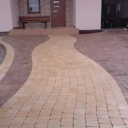 Ścieżka z kostki brukowej w dwóch odcieniach prowadząca do domu z drewnianą ławką przed wejściem.