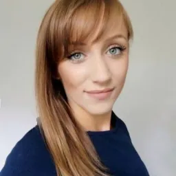 Portret uśmiechniętej kobiety z długimi, prostymi, brązowymi włosami z grzywką i niebieskimi oczami, ubrana w ciemny sweter.