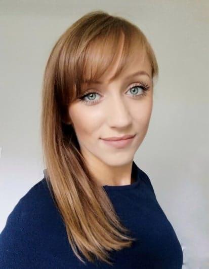 Portret uśmiechniętej kobiety z długimi, prostymi, brązowymi włosami z grzywką i niebieskimi oczami, ubrana w ciemny sweter.