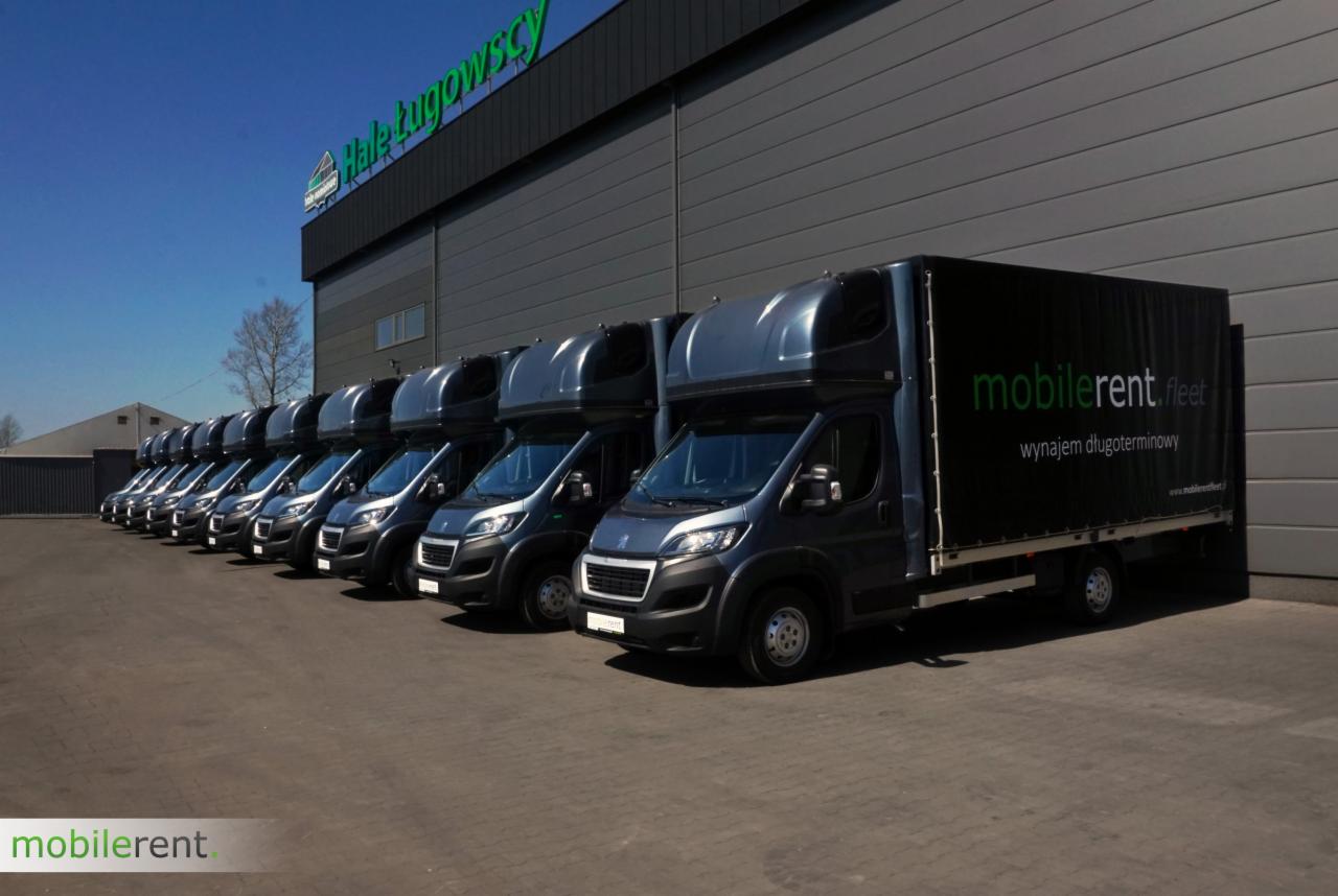 Flota szarych samochodów dostawczych z plandekami, ustawionych w rzędzie przed szarym budynkiem z zielonym logo firmy.