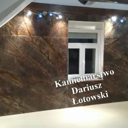 Ściana w pokoju na poddaszu wyłożona dużymi płytami z kamienia o wzorze przypominającym żyły drzew, oświetlona punktowymi lampkami, z oknem w białej ramie i fragmentem białej zabudowy kuchennej.