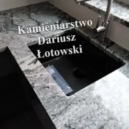 Szary blat kamienny z czarnym zlewem i nowoczesną baterią, widoczny fragment okna z naklejką reklamową Salamander, w zlewie przezroczysta torebka foliowa.