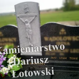 Granitowy pomnik z rzeźbą ukrzyżowanego Chrystusa, ozdobiony kwiatami, na tle zielonego krajobrazu.