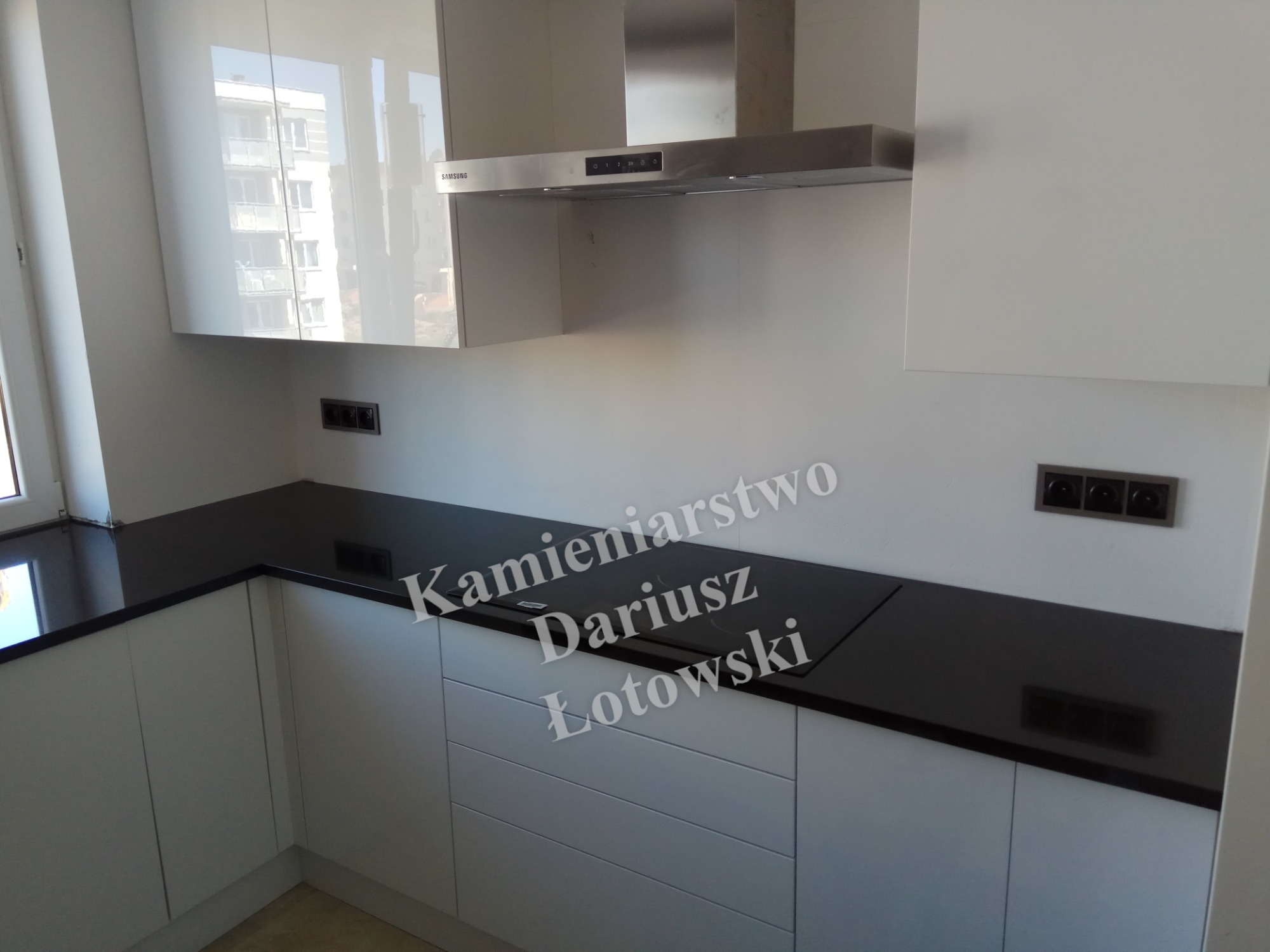 Nowoczesna kuchnia z czarnym blatem i białymi szafkami, widoczna płyta indukcyjna i okap Samsung, w tle budynek odbijający się w lakierowanych frontach szafek