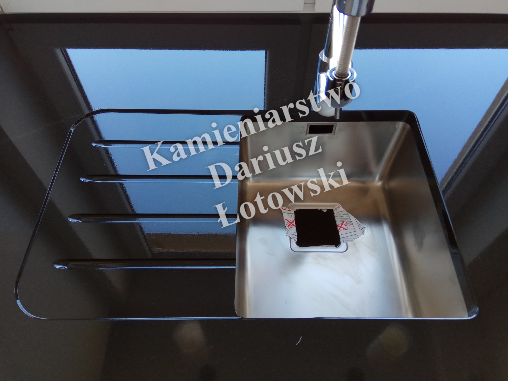 Błyszczący, czarny blat z kamienia z wbudowanym, metalowym zlewem kuchennym i baterią, z naklejką ochronną na dnie zlewu.
