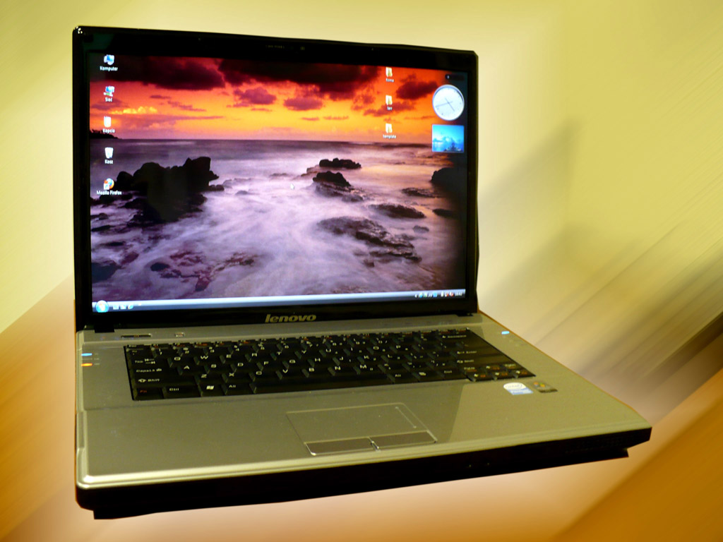 Laptop Lenovo z wyświetlonym pulpitem z motywem zachodu słońca nad morzem, ustawiony na biurku.
