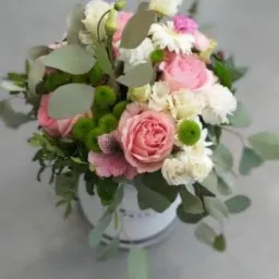 Kompozycja kwiatowa w okrągłym, białym pudełku z różami, goździkami, eustomą i zielonymi chryzantemami oraz eukaliptusem, na jednolitym, szarym tle.