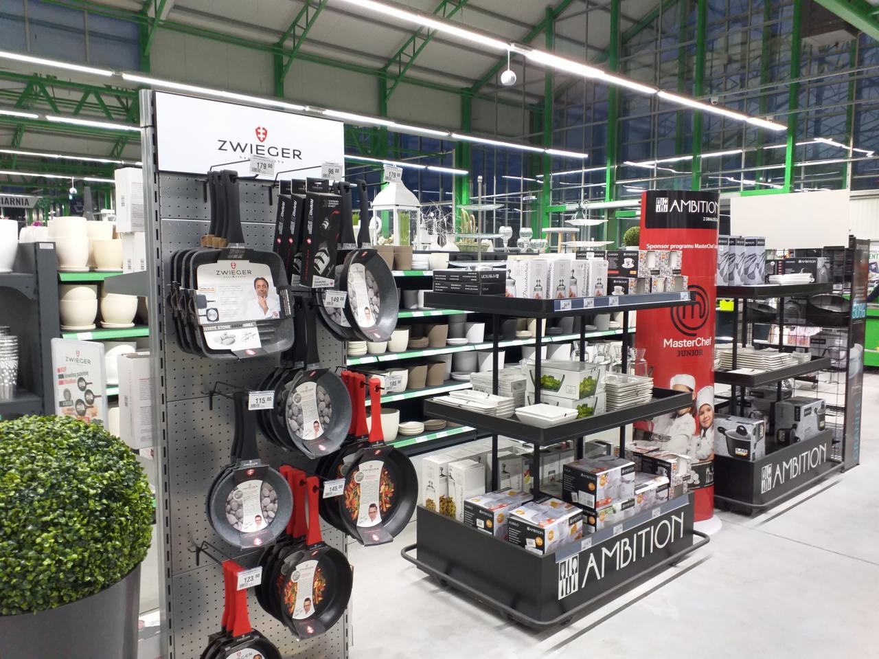 Ekspozycja sklepowa z różnorodnymi patelniami marki Zwiger i Ambition, naczyniami oraz akcesoriami kuchennymi na metalowych regałach w hali o konstrukcji stalowej.