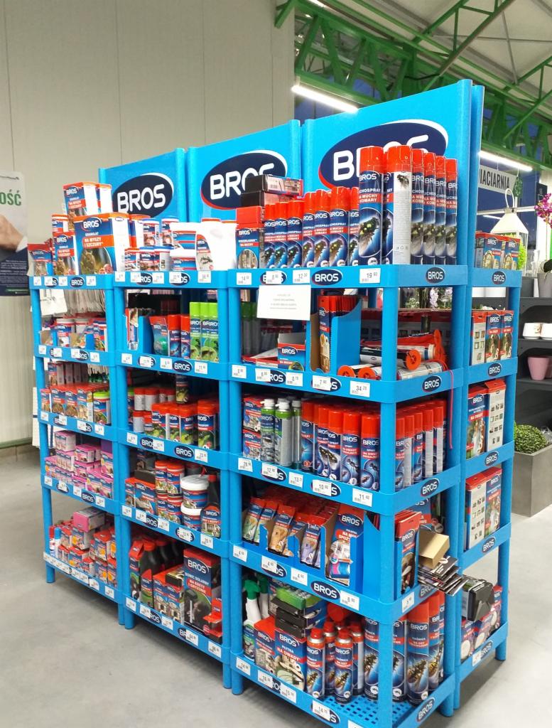 Niebieski regał sklepowy wypełniony produktami marki BROS, w tym sprayami i opakowaniami z grafiką zwierząt, na tle jasnej podłogi i fragmentu sufitu sklepu.