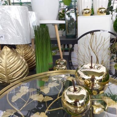 Złote dekoracje w kształcie jabłek i liści na szklanym stoliku z motywem liści, w tle lampa z białym abażurem i wysokie zielone trawy w doniczce, całość w jasnej, słonecznej przestrzeni sklepu.