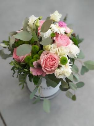 Kompozycja kwiatowa w okrągłym, białym pudełku z różami, goździkami, eustomą i zielonymi chryzantemami oraz eukaliptusem, na jednolitym, szarym tle.