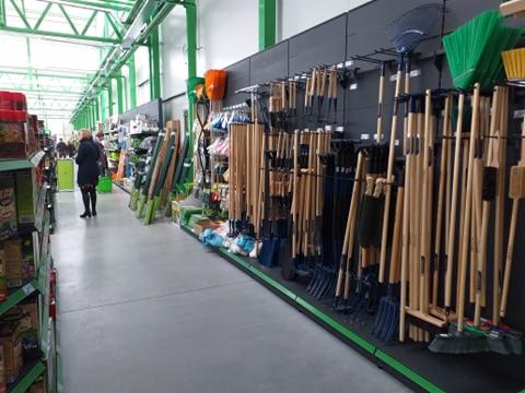 Wnętrze sklepu ogrodniczego: Rząd narzędzi ogrodowych z drewnianymi trzonkami i kolorowymi końcówkami (łopaty, grabie, widły) zawieszonych na czarnej ścianie z zielonymi elementami konstrukcyjnymi.