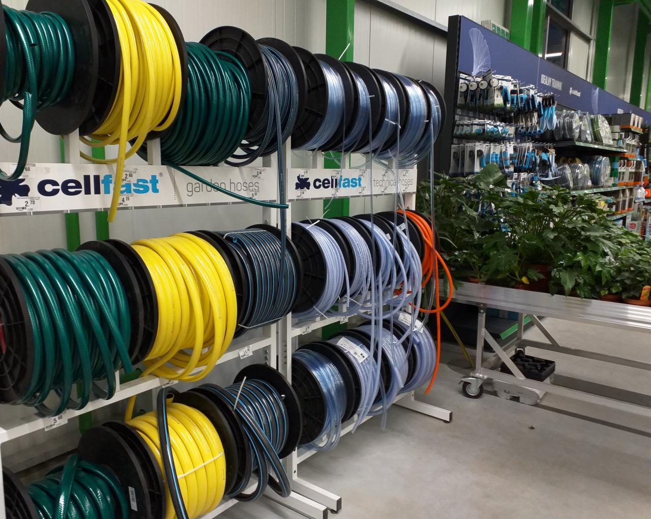 Regał sklepowy z nawiniętymi wężami ogrodowymi w kolorach zielonym, żółtym, niebieskim i pomarańczowym, z logo 'cellfast' i roślinami doniczkowymi w tle.