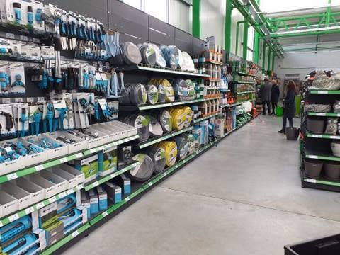Wnętrze sklepu ogrodniczego z półkami wypełnionymi narzędziami, wężami ogrodowymi, zraszaczami i podłożami. Widoczni klienci w tle.