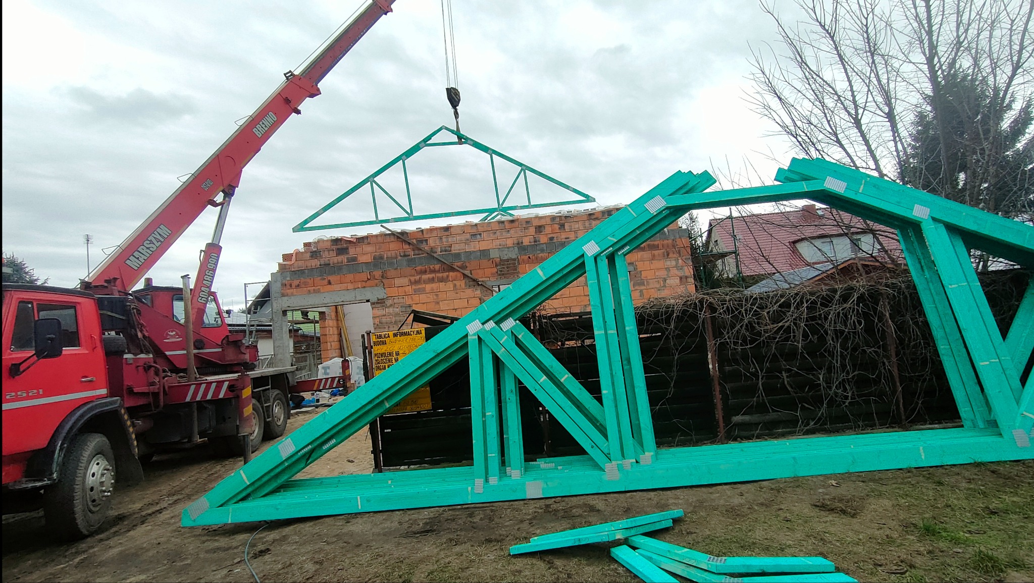 Montaż zielonej więźby dachowej z użyciem czerwonego dźwigu Marszym na tle murowanego budynku w trakcie budowy.