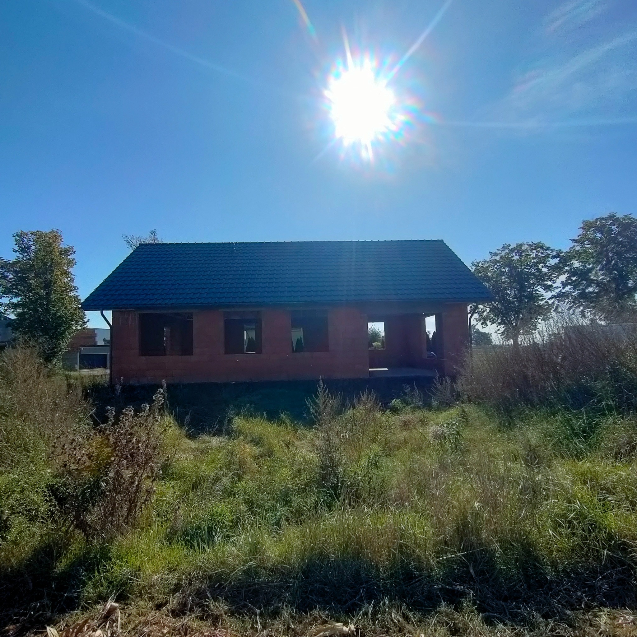 Niedokończony budynek w stanie surowym otwartym z ciemnym dachem z dachówki, widoczny na tle jasnego, słonecznego nieba i zarośniętego terenu.