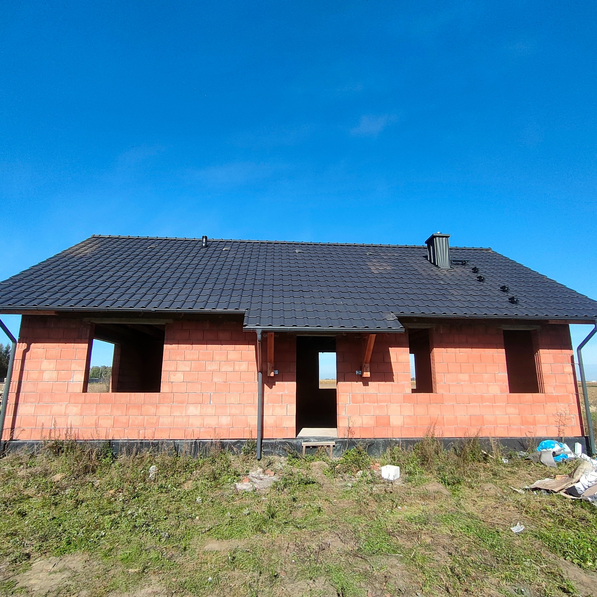 Niedokończony dom z cegły z czarnym dachem z dachówki ceramicznej i zamontowanymi rynnami, widoczne otwory okienne i drzwiowe, na tle błękitnego nieba.