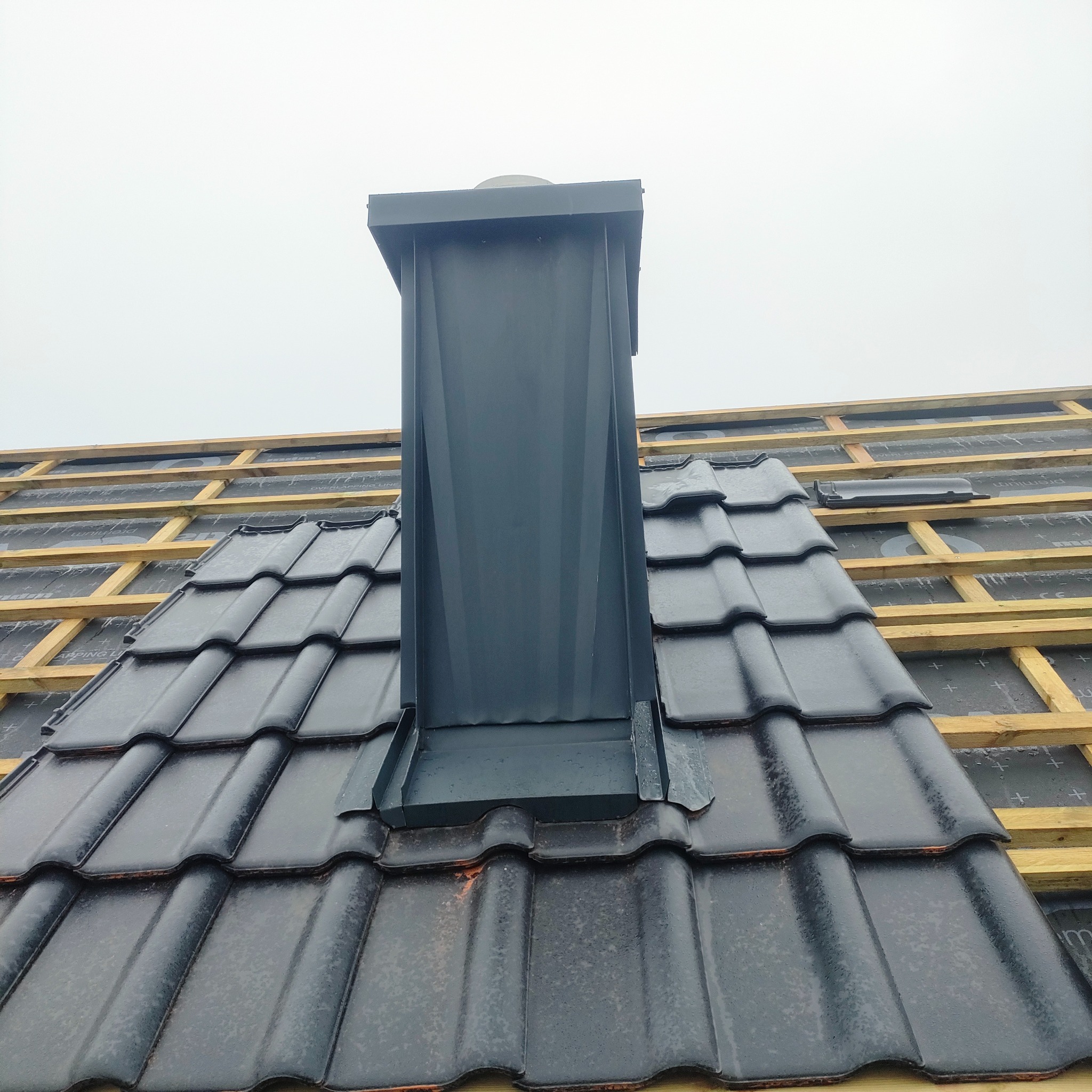 Nowe, błyszczące dachówki ceramiczne na dachu z kominem w trakcie montażu, widoczne drewniane łaty i folia dachowa w tle, pochmurne niebo.