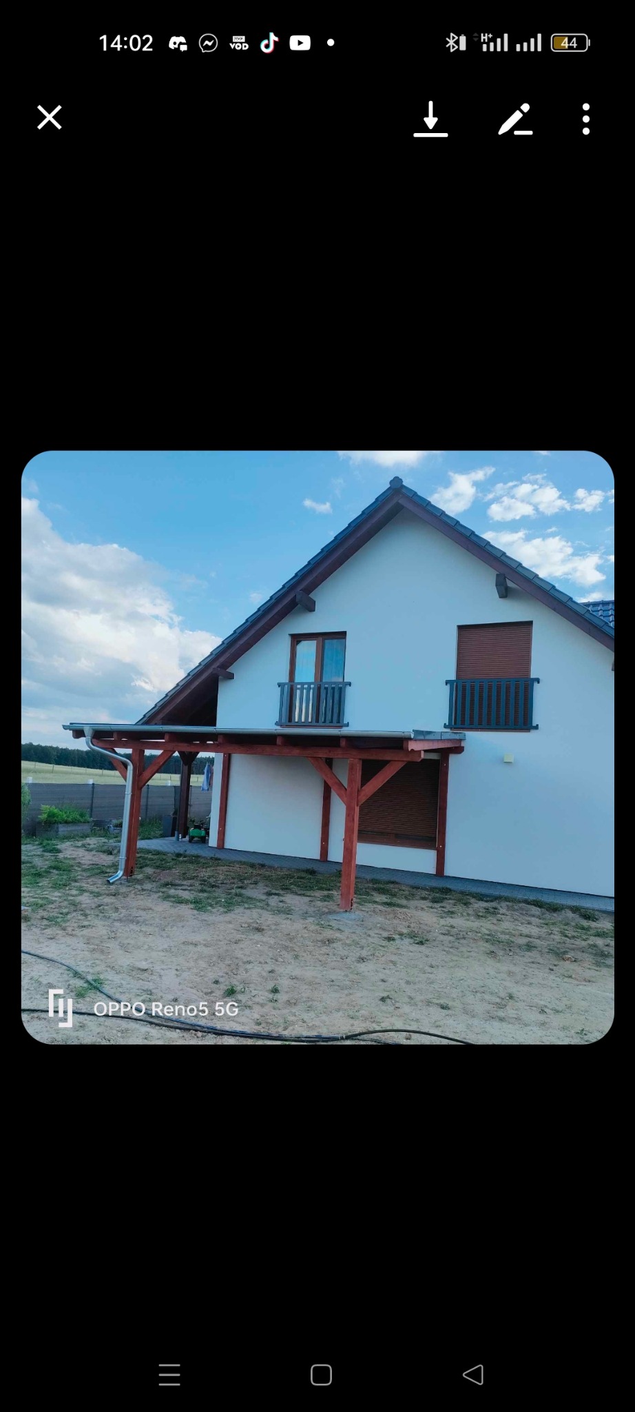 Nowoczesny dom z drewnianą konstrukcją zadaszenia tarasu, widoczne brązowe słupy i beżowa elewacja budynku, w tle błękitne niebo z chmurami.