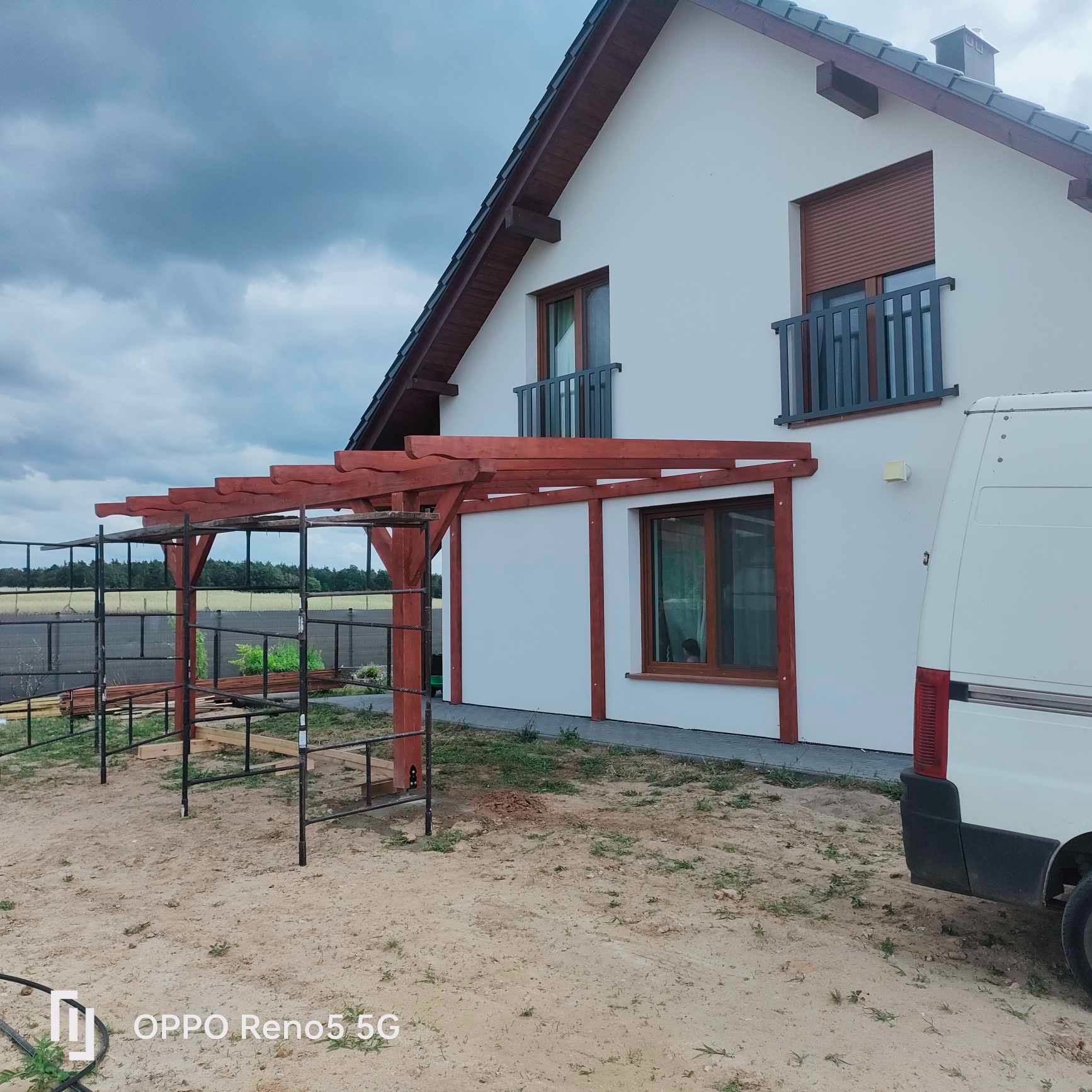 Montaż drewnianej konstrukcji przy białym domu z brązowym dachem, widoczne rusztowania i częściowo zaparkowany biały samochód dostawczy.