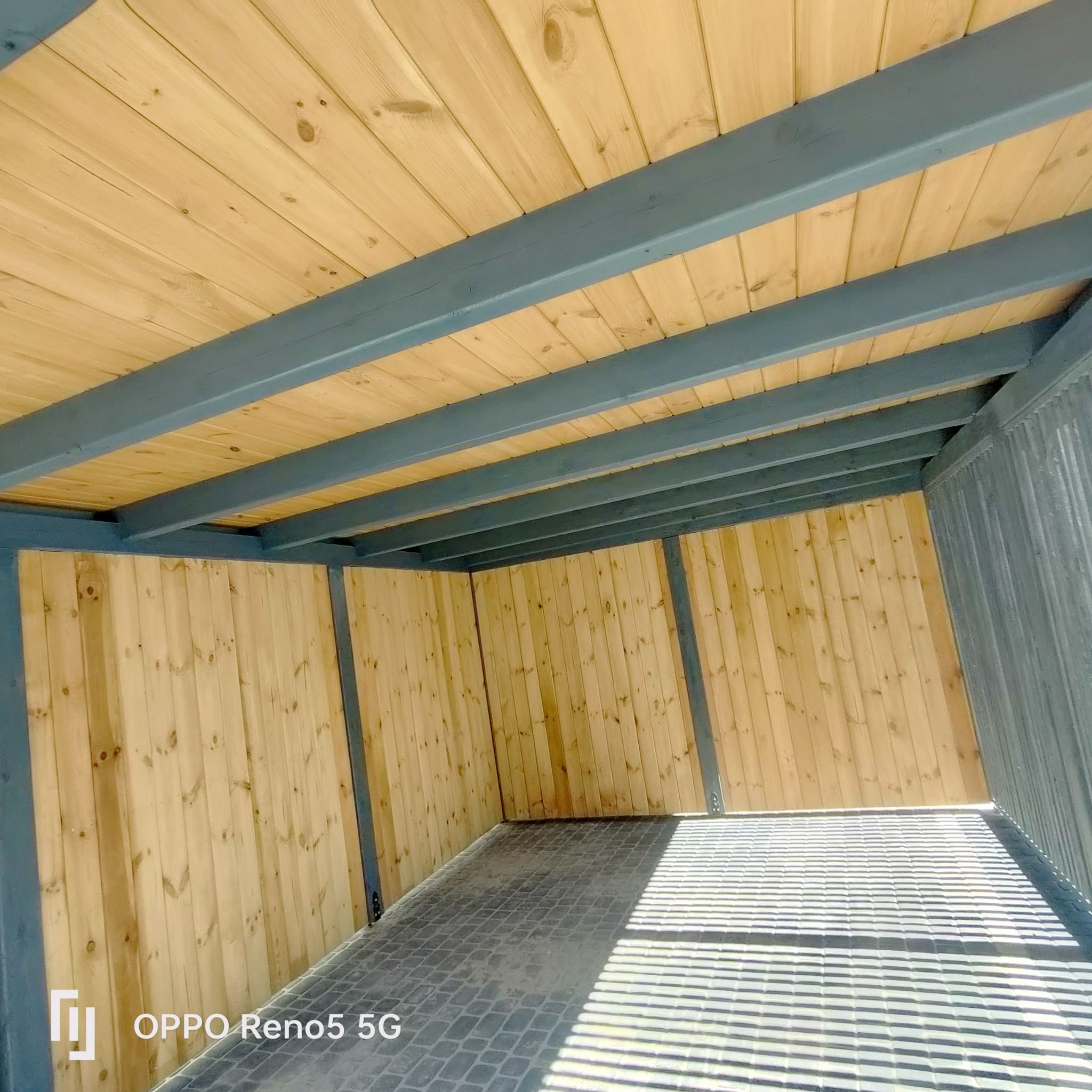 Wnętrze drewnianej konstrukcji z szarą ramą, jasne drewniane deski ścian i sufitu, ciemna kostka brukowa na podłodze, widok z niskiej perspektywy.