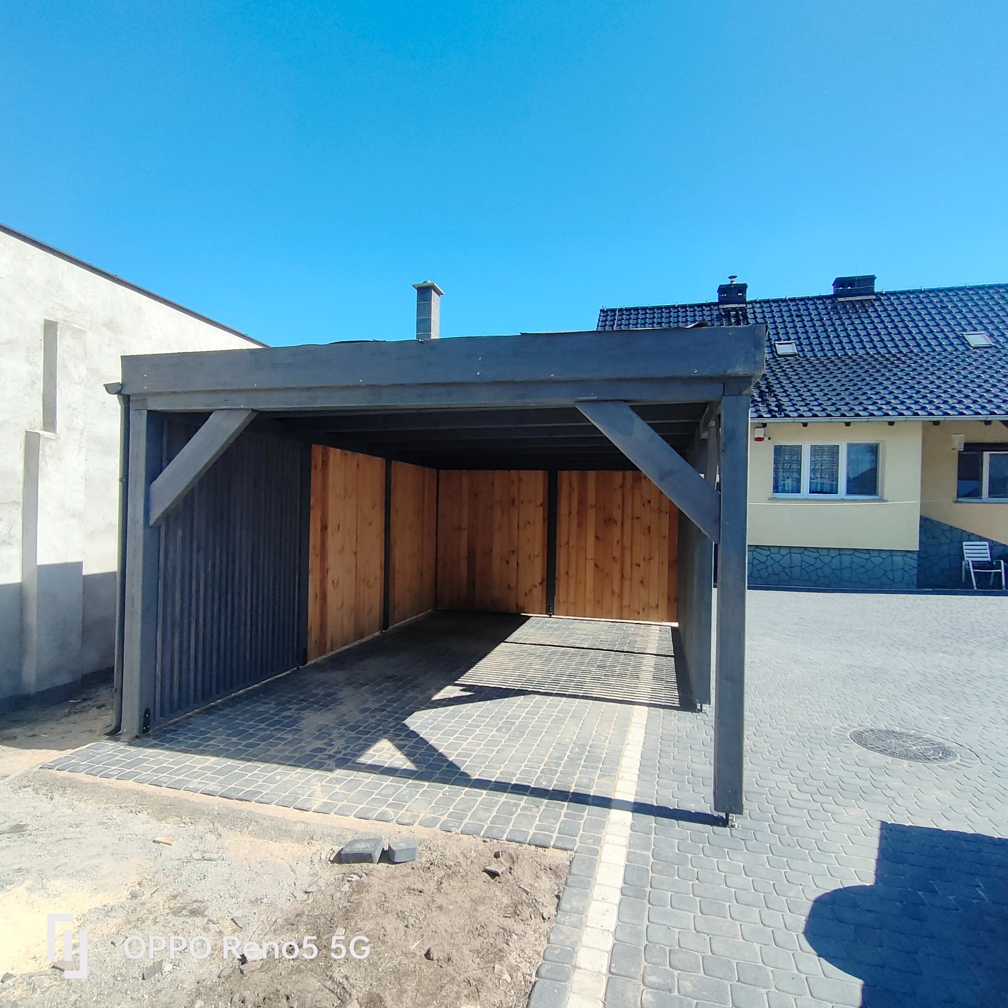 Ciemnoszara pergola z drewnianym wypełnieniem ścian bocznych, z kostki brukowej ułożony podjazd, w tle dom z ciemnym dachem i jasną elewacją.