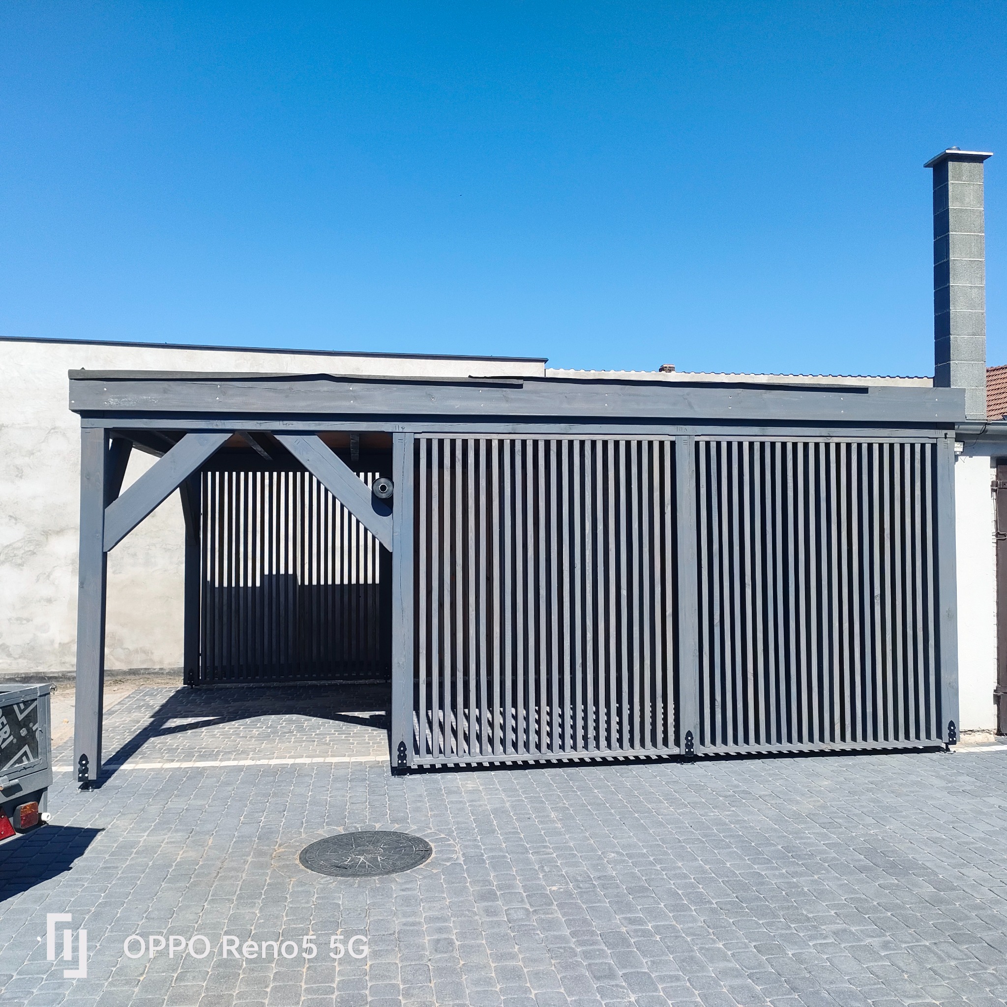 Nowoczesna, szara pergola z drewnianymi lamelami, częściowo zasłonięta, na posesji we Włoszakowicach, z kostką brukową i błękitnym niebem w tle.