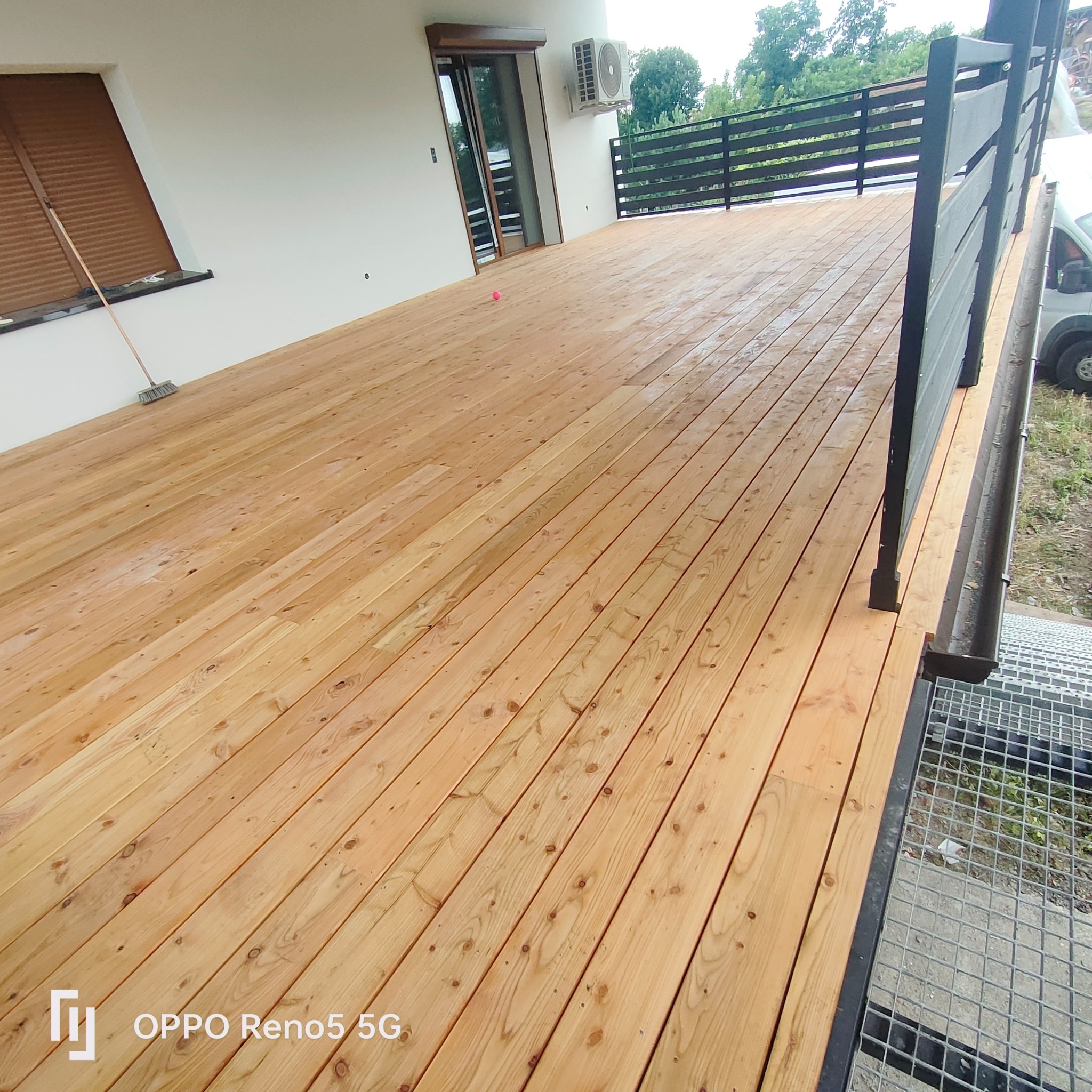 Nowo wybudowany taras z jasnego drewna, z czarną balustradą i widocznym fragmentem schodów z metalowej siatki.