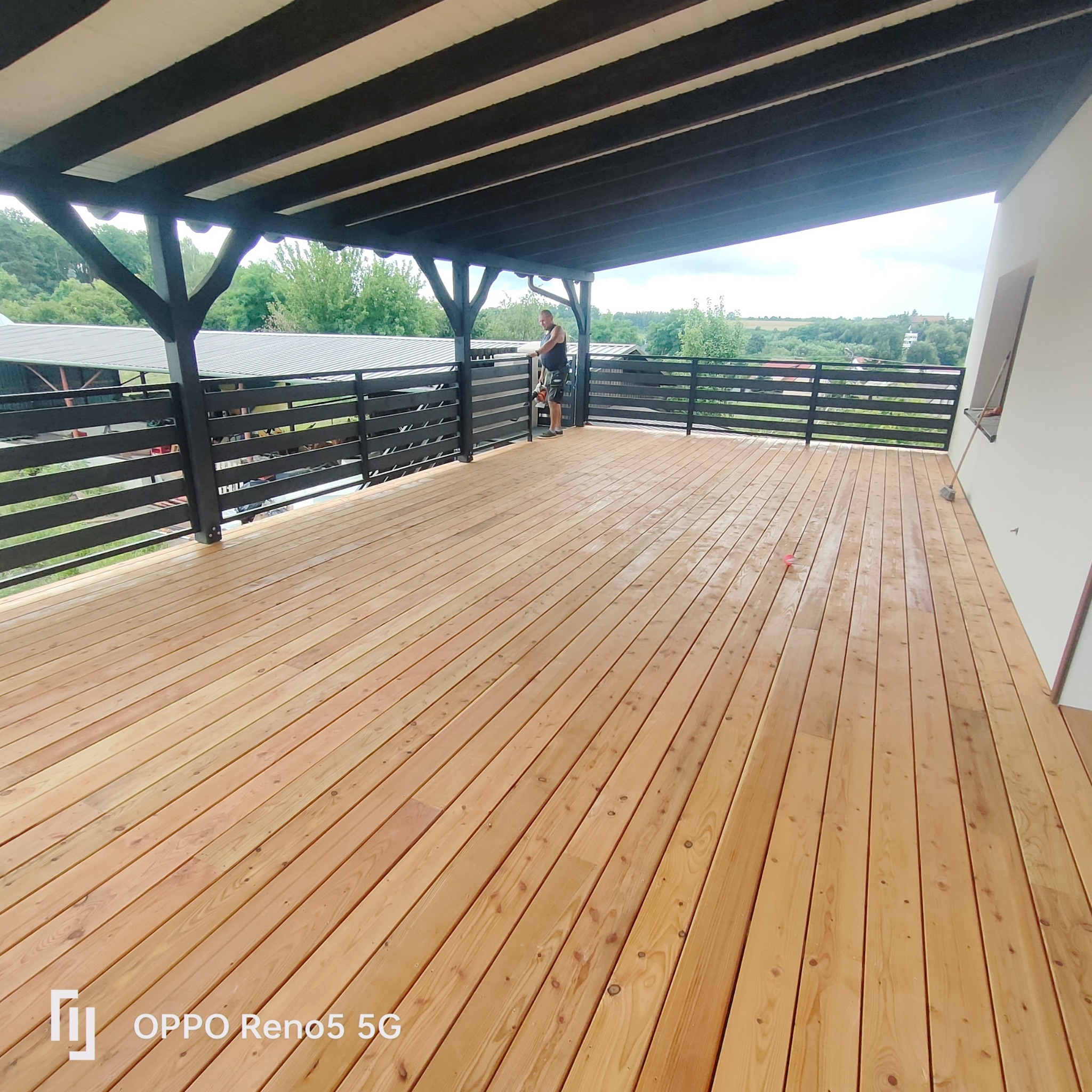 Świeżo wybudowany drewniany taras z czarną balustradą i zadaszeniem, widok na zielony krajobraz w tle, pracownik z narzędziem przy balustradzie.