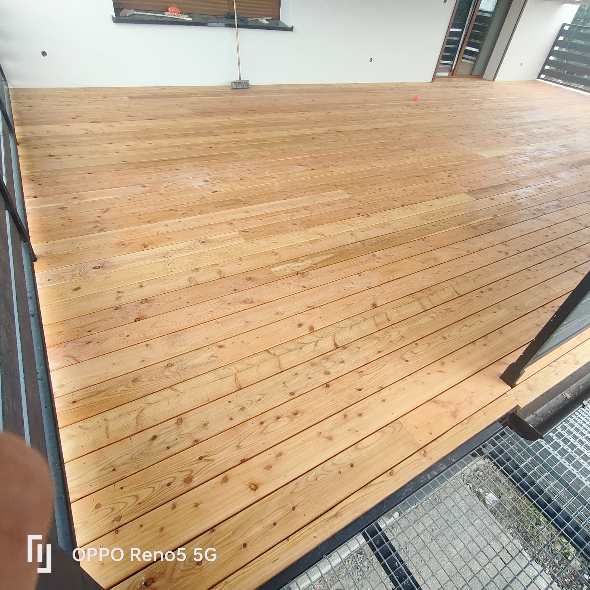 Nowo wybudowany, jasny taras z desek drewnianych, widok z góry, z widocznym fragmentem balustrady i ścianą budynku w tle, z pozostawioną miotłą.