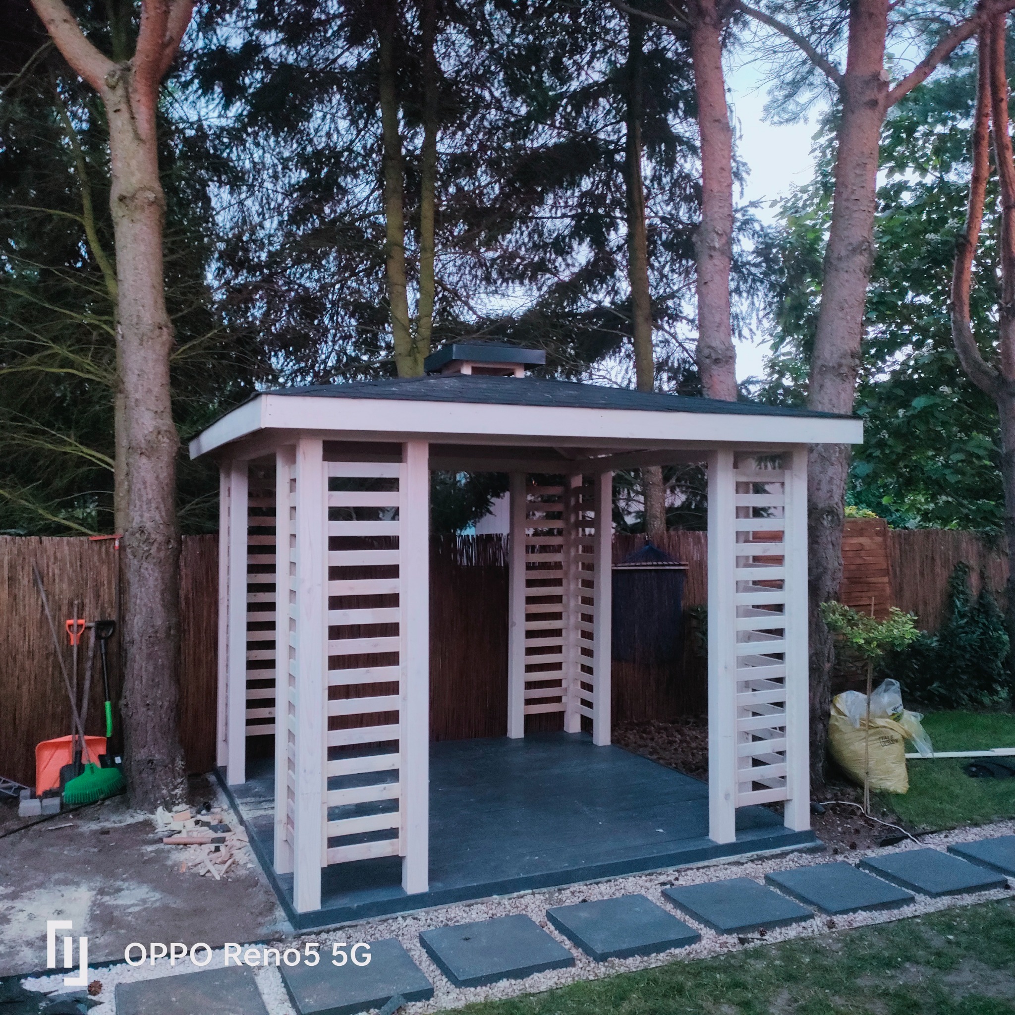 Nowa, jasna konstrukcja z drewna z ażurowymi ścianami w ogrodzie, z widocznymi narzędziami i śladami pracy w tle drzew i bambusowego płotu.