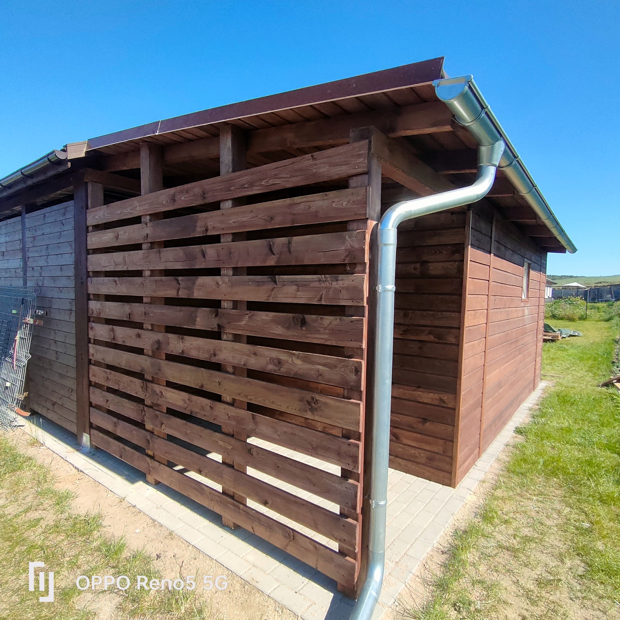 Drewniana altana z poziomym ażurowym panelem bocznym, brązowe deski, metalowa rynna, widok z zewnątrz w słoneczny dzień.