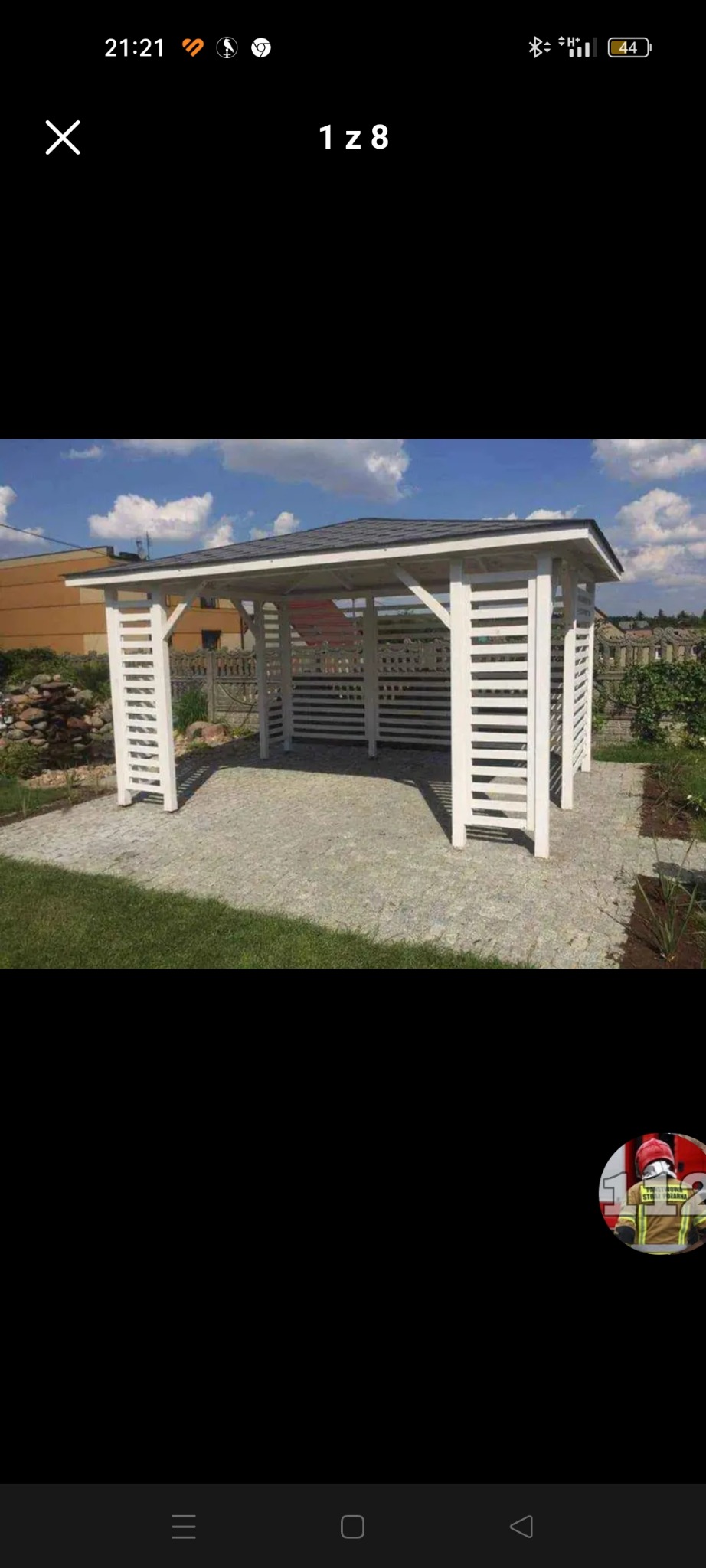 Biała, drewniana konstrukcja z dachem z gontu bitumicznego, przypominająca pawilon ogrodowy, ustawiona na brukowanej powierzchni, z widocznym trawnikiem i budynkami w tle.