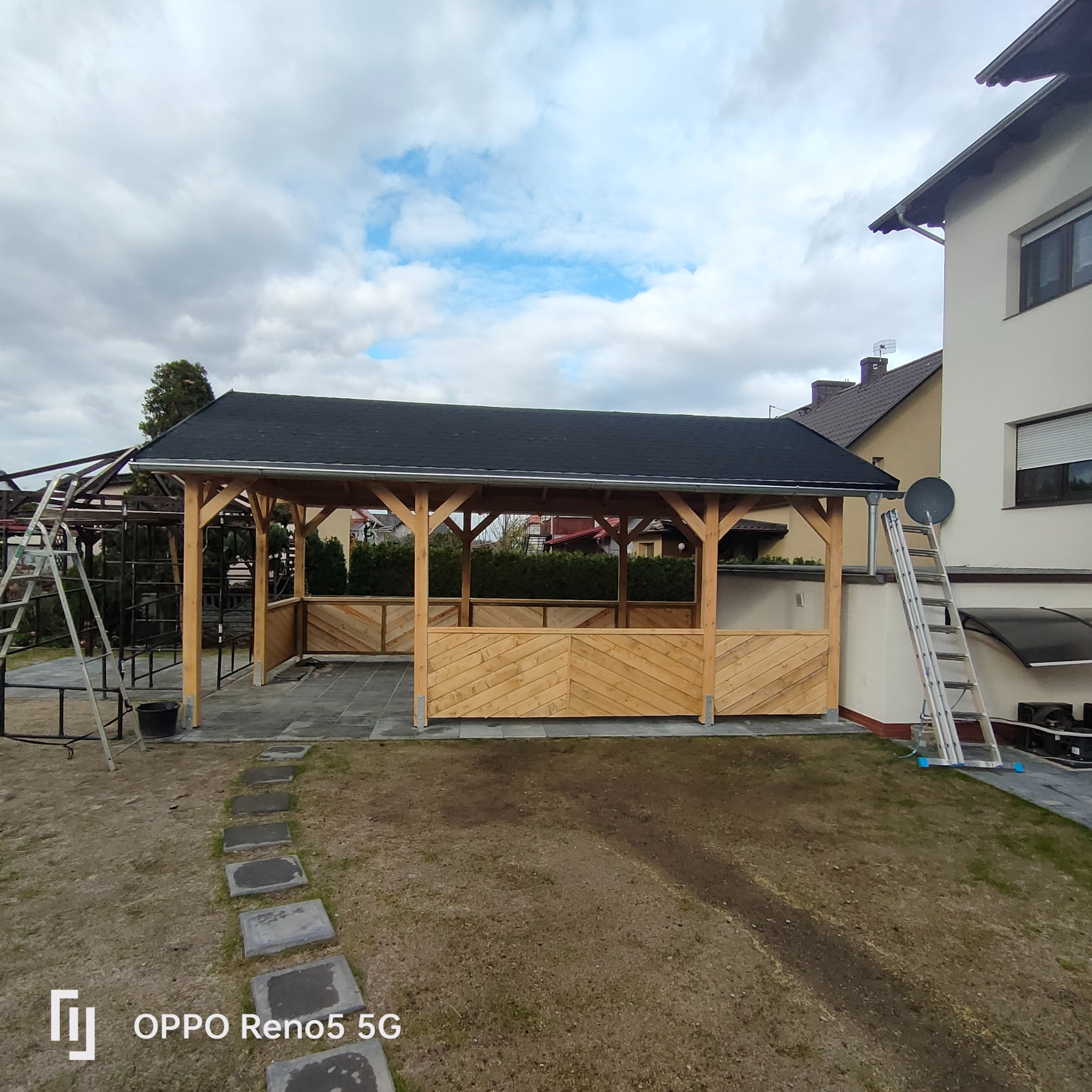 Surowa drewniana konstrukcja z czarnym dachem, częściowo osłonięta ukośnymi deskami, widoczna na tle domu i pochmurnego nieba, z elementami rusztowania i drabiną obok.