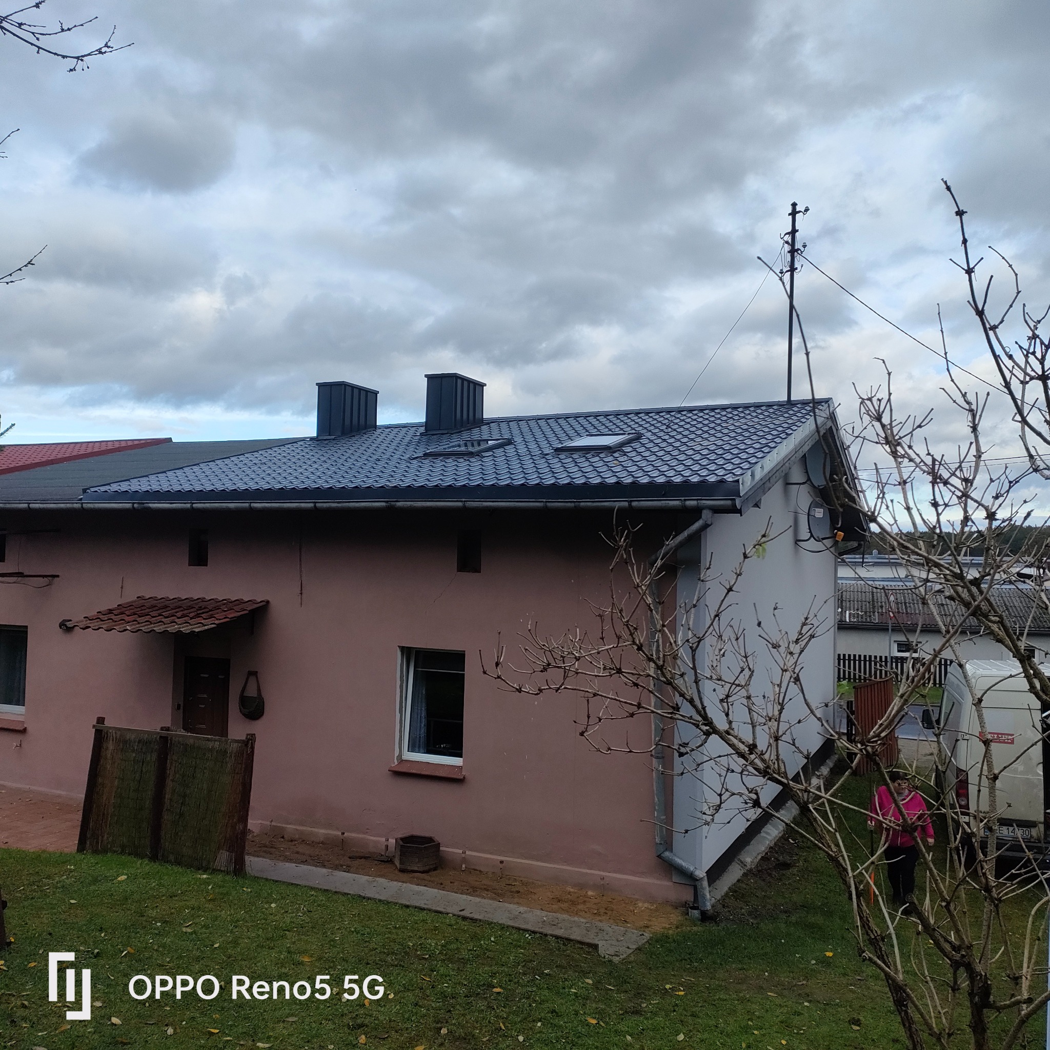 Dom z nowym, ciemnym dachem z blachodachówki, widoczne dwa kominy i okna dachowe, na ścianie budynku satelita, w tle pochmurne niebo.