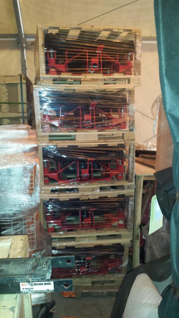 Części do wózków widłowych w drewnianych skrzyniach na paletach, zabezpieczone folią, składowane w magazynie w Kornicy. Widoczna czarna belka z naklejką 'Nasadka wideł'.