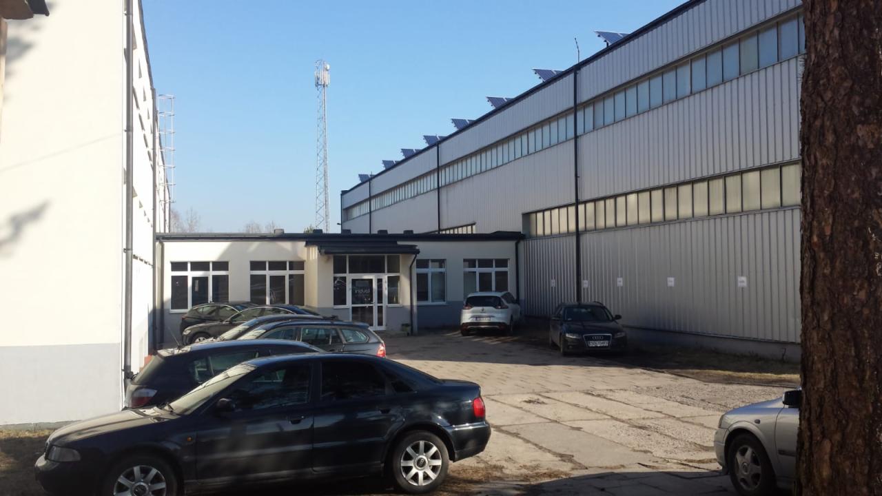 Widok z zewnątrz na magazyn z panelami słonecznymi na dachu, zaparkowanymi samochodami i wieżą telekomunikacyjną w tle.