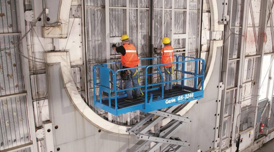 Dwóch pracowników w pomarańczowych kamizelkach i żółtych kaskach na niebieskim podnośniku nożycowym Genie GS-3246, wykonujących prace na dużej, metalowej konstrukcji z widocznymi śrubami i panelami.