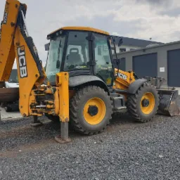 Żółto-czarna koparko-ładowarka JCB 4CXeco zaparkowana na żwirowym placu, z podniesionym ramieniem koparki i opuszczonymi podporami, na tle szarego budynku z bramami garażowymi.